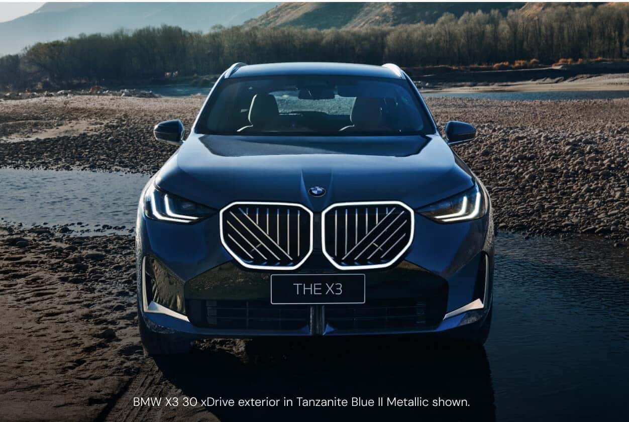 Front view of a BMW X3 30 xDrive in Tanzanite Blue showcasing its illuminated kidney grille on a rocky riverbed.