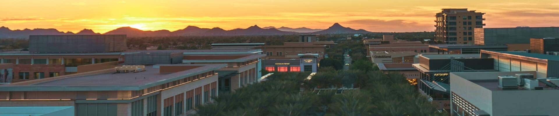 City shopping district at sunset, with modern buildings, palm trees and distant mountain silhouettes.