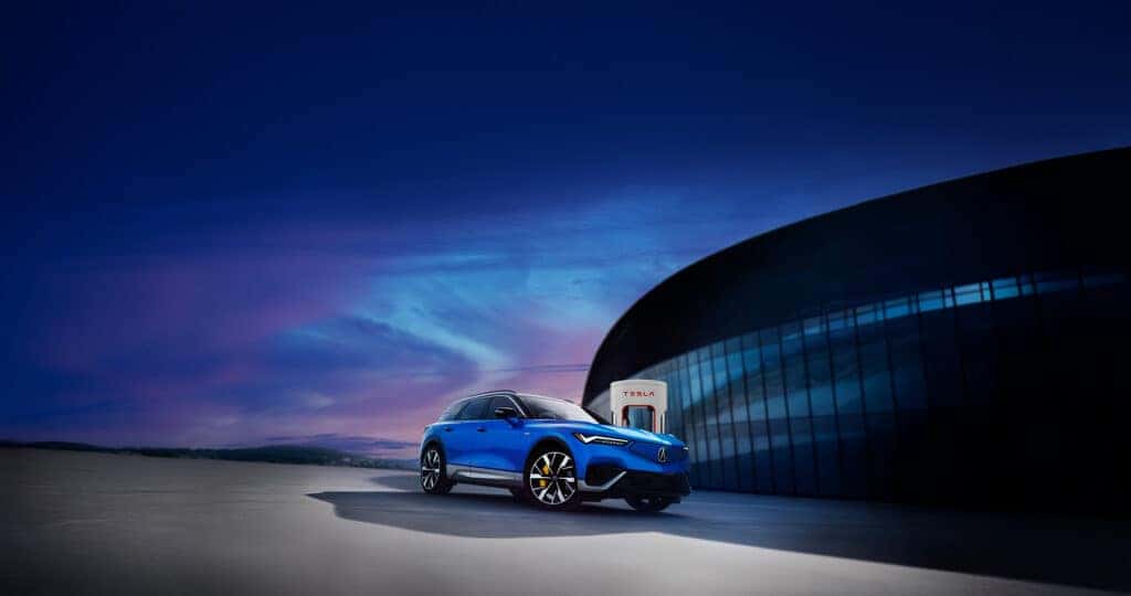 Charging the Acura ZDX at Tesla Supercharger in a parking lot outside of a large glass building at sunset