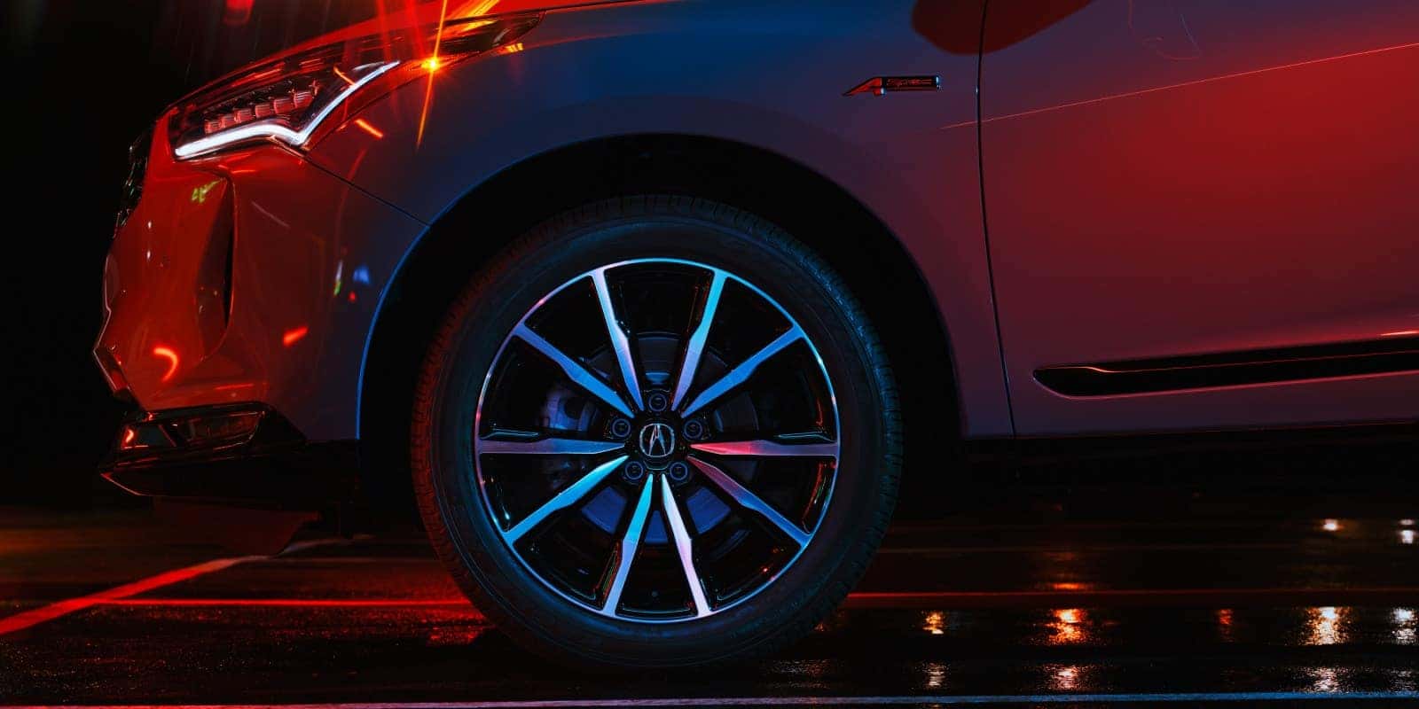Close-up of the 2026 Acura RDX A-Spec front wheel, LED headlight, and sculpted front fender under dramatic red and blue studio lighting.