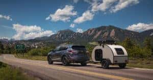 2026 Acura RDX towing a small teardrop camper along a scenic road outside Boulder, with the Flatirons rising in the background and Colorado foothills stretching beneath a bright blue sky.
