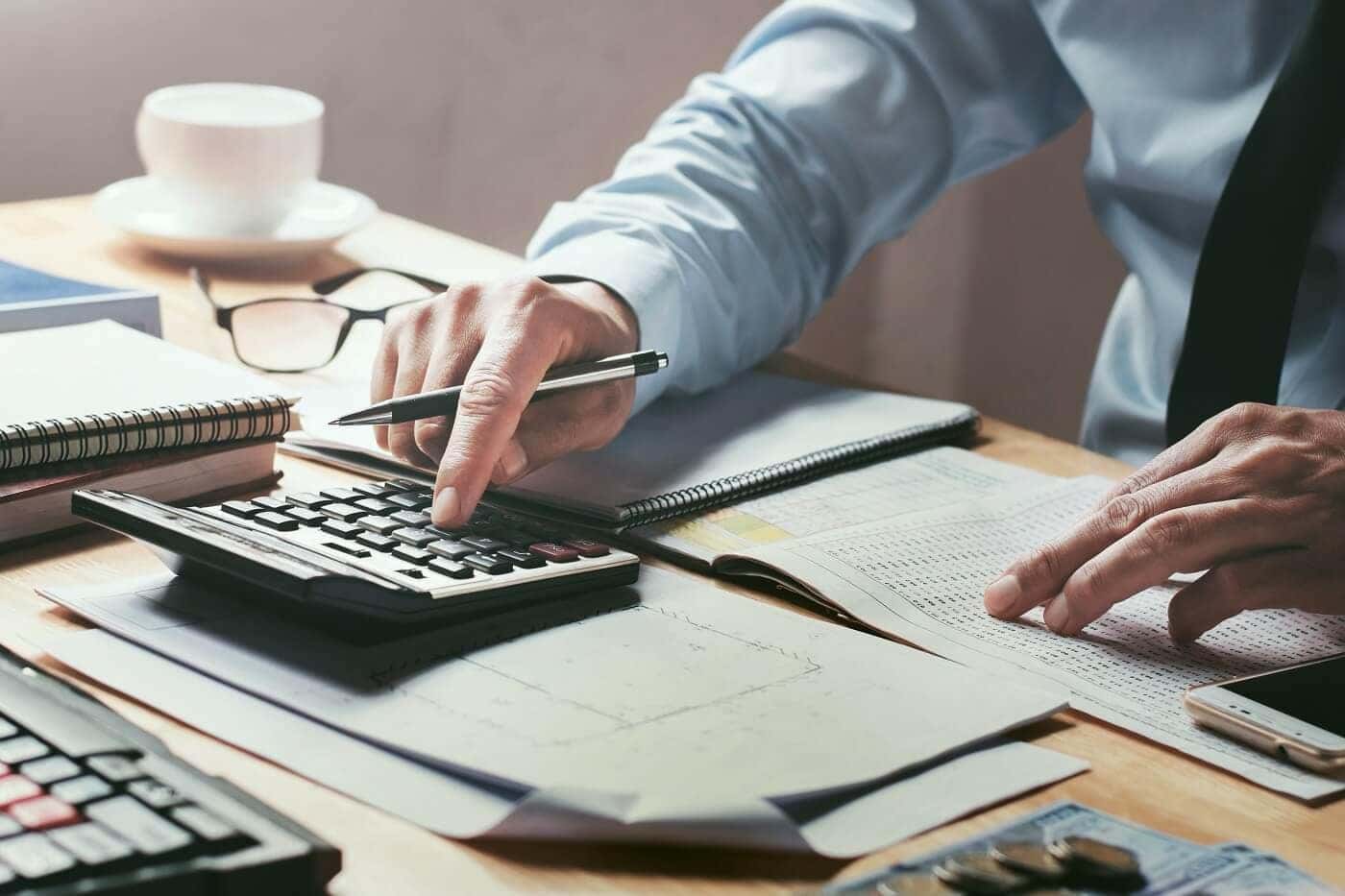 A hand presses a calculator that sits on a desk filled with paperwork.