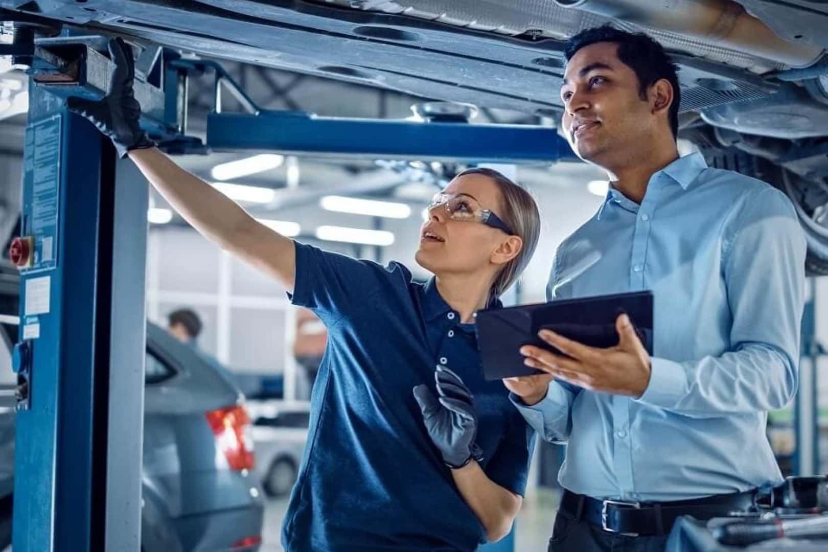 A female mechanic points up towards a vehicle as a male technician holds a table.