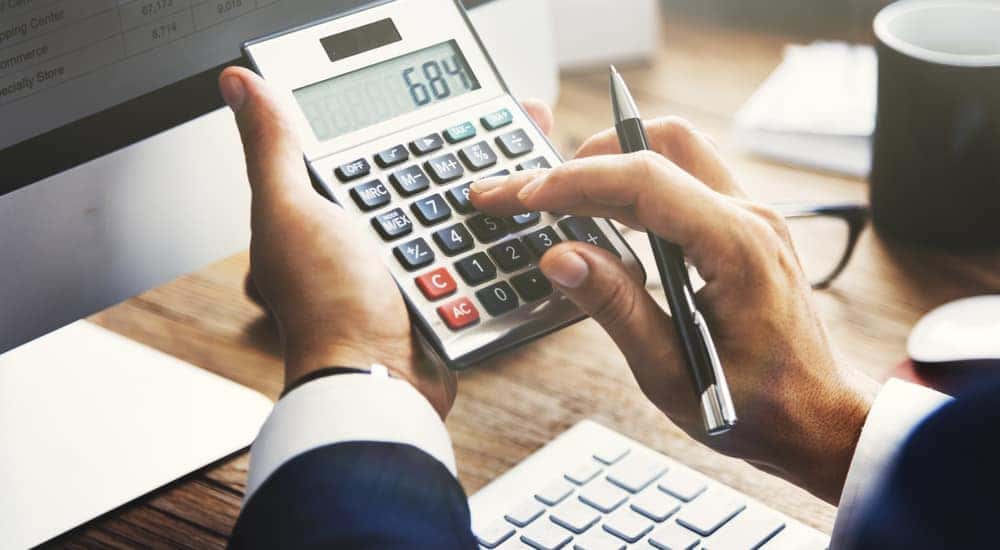 Hands of a man in a suit holding a calculator in front of a computer on a brown desk