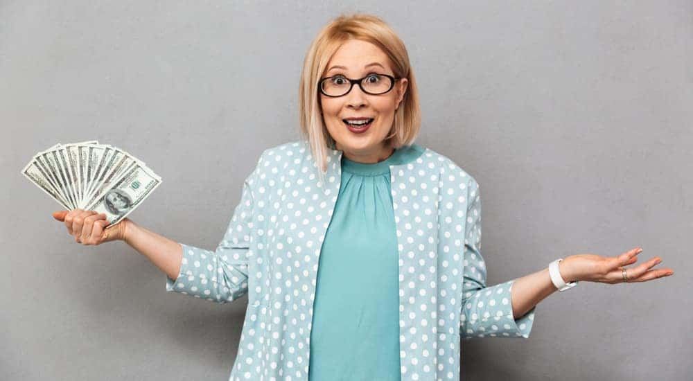 Blonde woman with glasses in a blue shirt and blue and white polka dot cardigan shrugging with cash