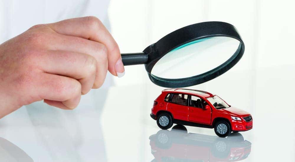 A hand holding a magnifying glass over a red toy car, reflective white background