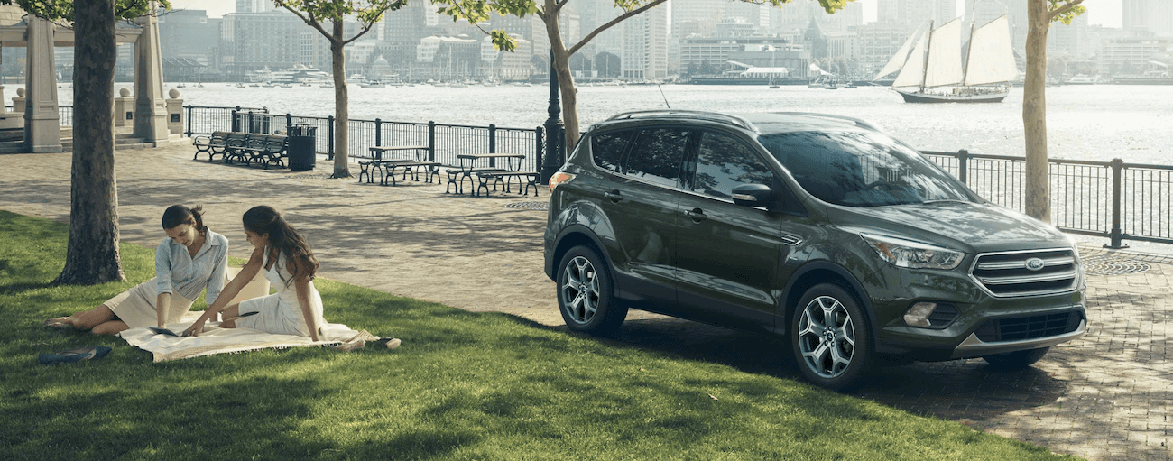 A green 2019 Ford Escape is parked next to a city park where two women are sitting on a blanket and a sailboat is in the water behind it.