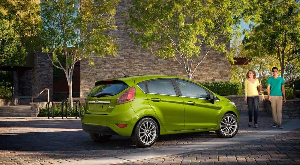 A couple walking past a green Ford Fiesta on a paver driveway with trees and a stone building in back