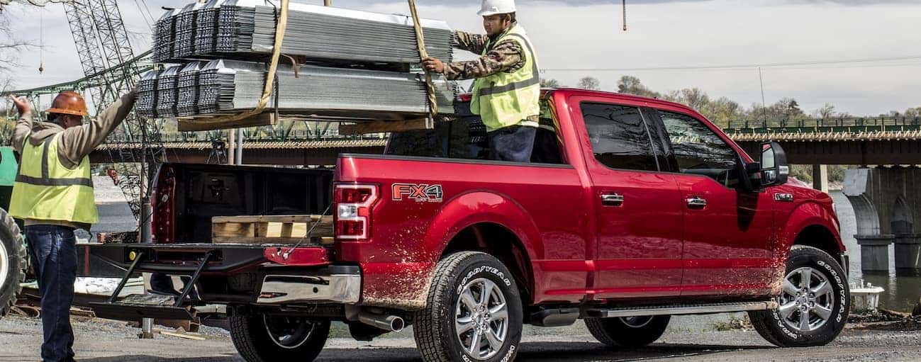 Two workers with yellow vests are putting heavy metal roofing material in the bed of a red 2020 Ford F-150.