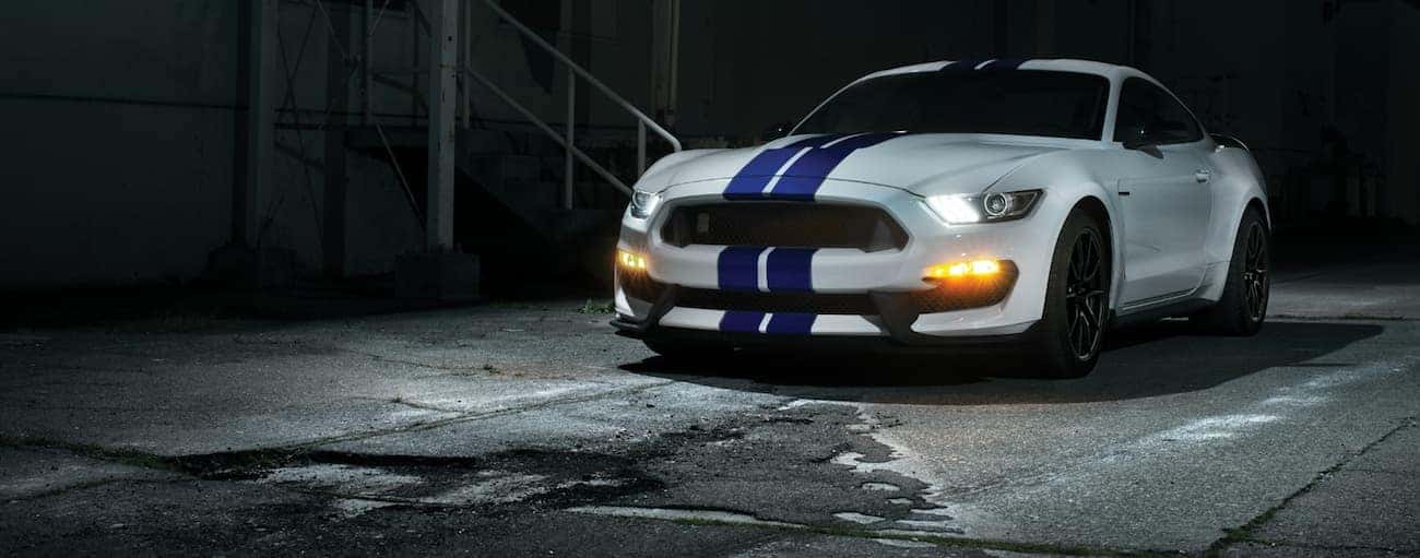 A white 2020 Ford Mustang Shelby GT350 with blue stripes is parked in a dark alleyway with its headlights on.