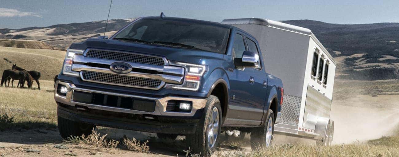 A blue 2020 Ford F-150 is towing a large horse trailer on a farm.