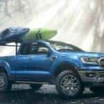 A blue 2020 Ford Ranger is parked on a wooded trail with kayaks on the bed racks.