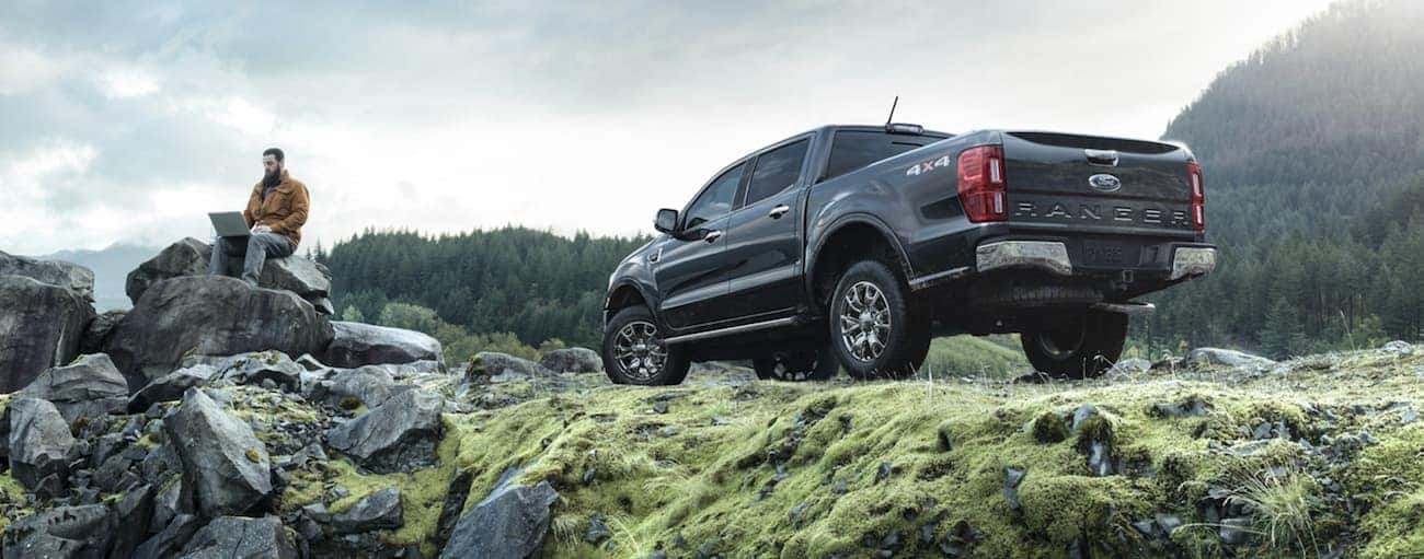 A man is sitting on a rock near his dark grey 2020 Ford Ranger parked on grass.