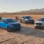 A blue, grey, and a silver 2020 Ford F-150 are parked in the desert with dirt bikes and side-by-sides.