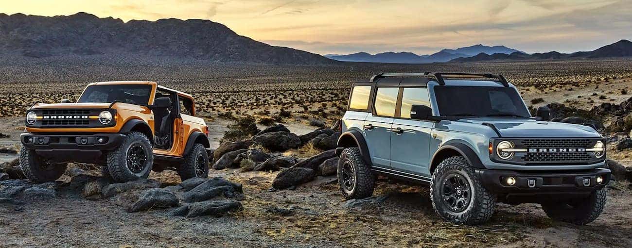 A yellow 2-Door and a blue 4-Door Ford Bronco are parked in the desert in front of mountains.