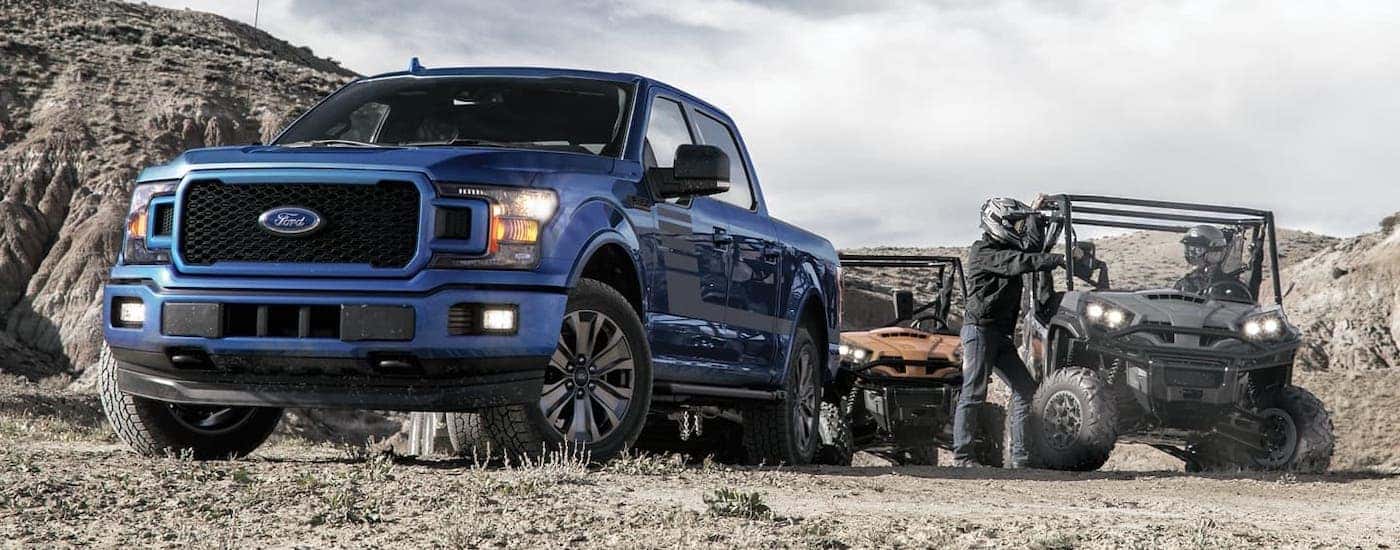 Two people are next to a blue 2020 Ford F-150 with 2 Side by Sides on a mountain trail.
