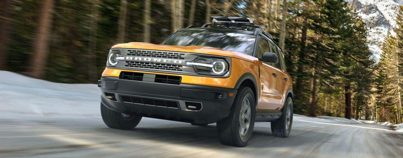 A yellow 2021 Ford Bronco Sport is driving on a snow-lined road past large trees.