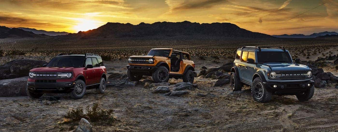 A red 2021 Ford Bronco Sport and an orange 2021 Ford Bronco 2-door and blue 4-door are parked on rocks in front of a desert sunset.