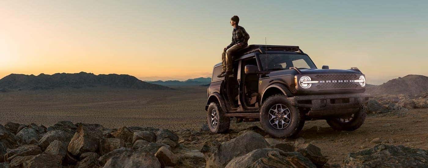 A man is sitting on the roof of a gray 2021 Ford Bronco four-door with no doors and overlooking a desert.