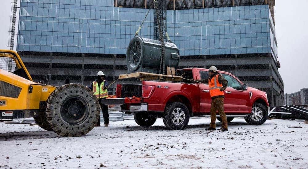 Workers are loading machinery into the bed of a red 2021 Ford F-150 in the snow.