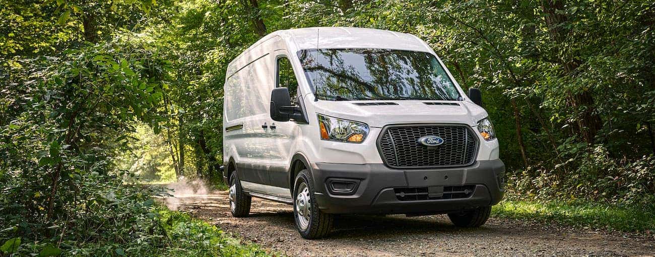 A white 2021 Ford Transit van is driving on a dirt road in the woods.