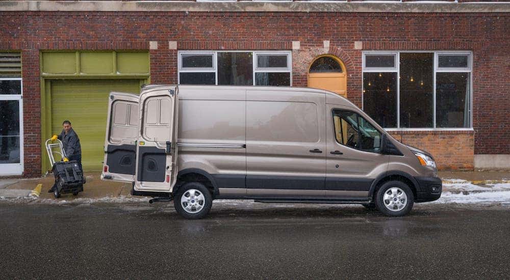 A man is loading cargo into the rear of a popular Ford commercial vehicle, a brown 2021 Ford Transit cargo van, in front of a brick building.