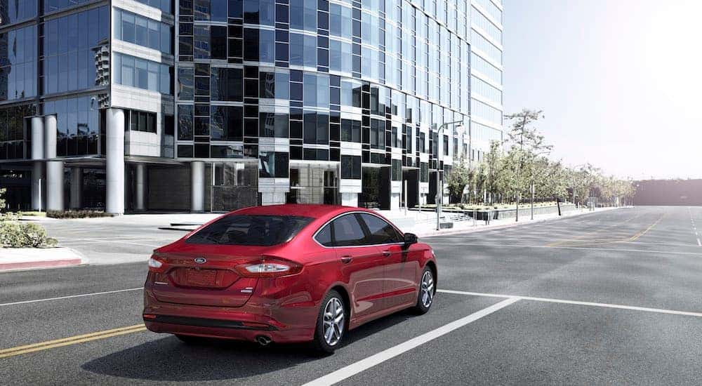 A red 2016 Ford Fusion is shown from behind parked at an intersection with a glass building in the background.