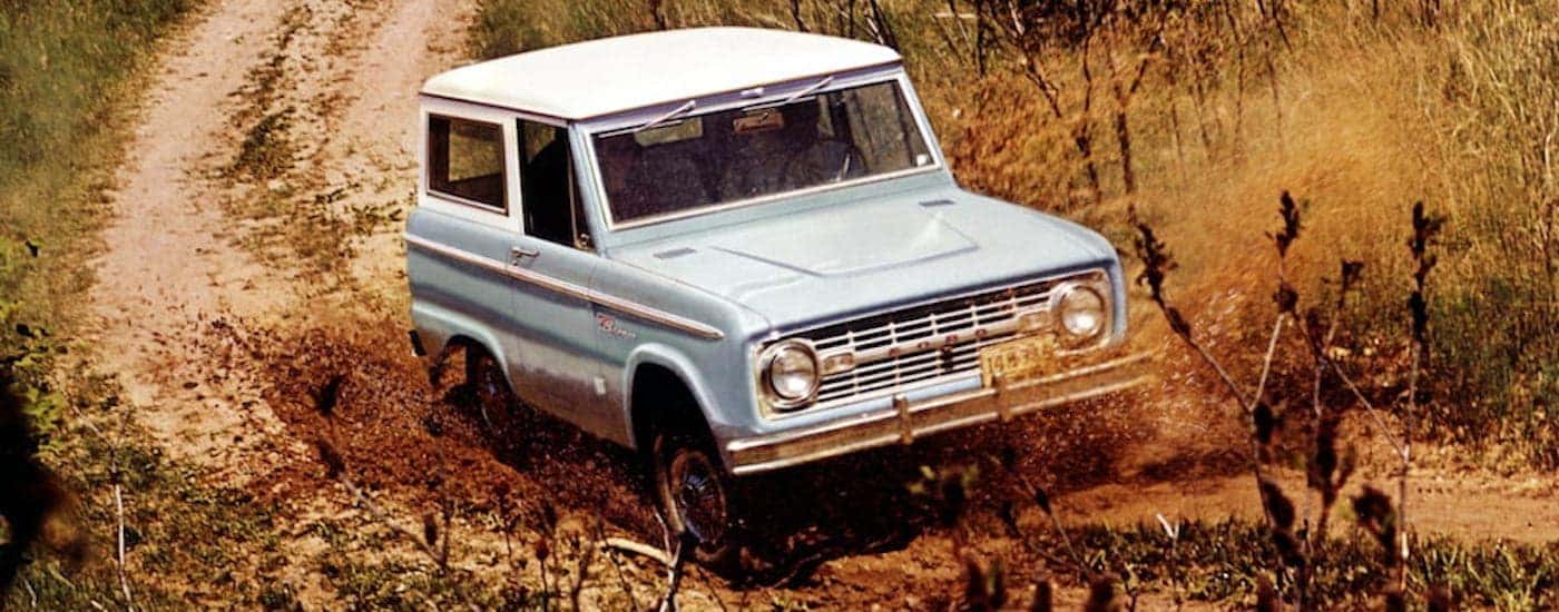A light blue 1967 Ford Bronco is off-roading on a muddy dirt trail.