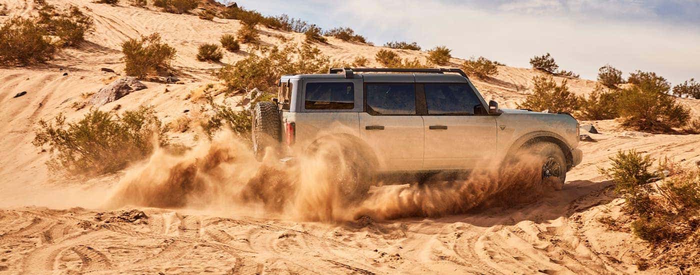 A silver 2021 Ford Bronco 4 door is shown from the side kicking up sand on a dune.