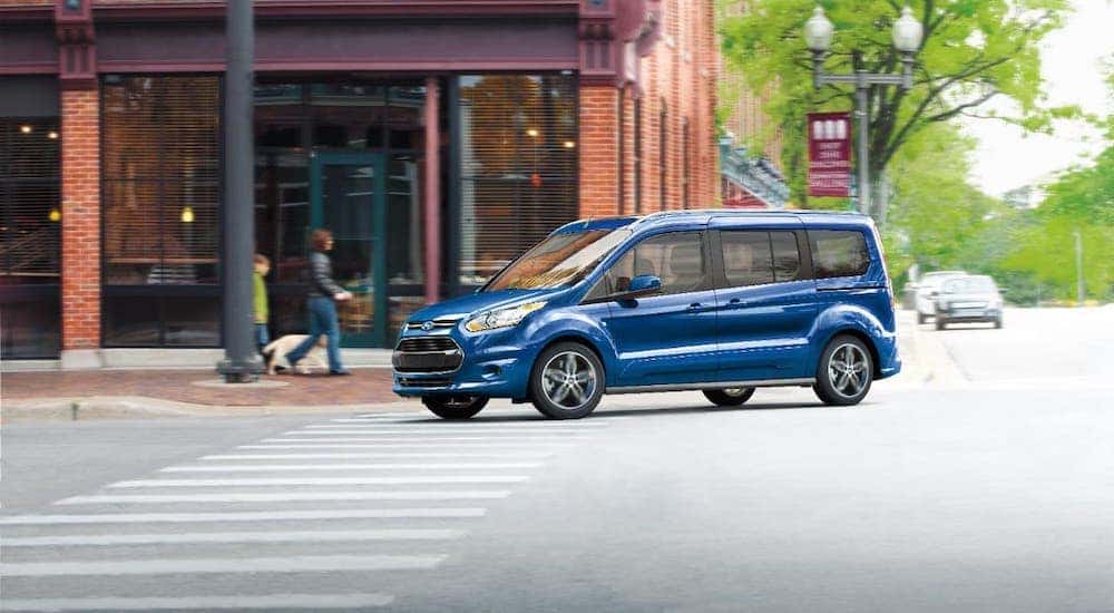 A blue 2019 Ford Transit Connect is driving around a corner in front of a red brick building.