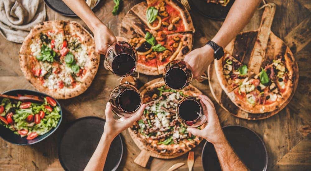 A top-down view shows people toasting wine glasses over several pizzas.