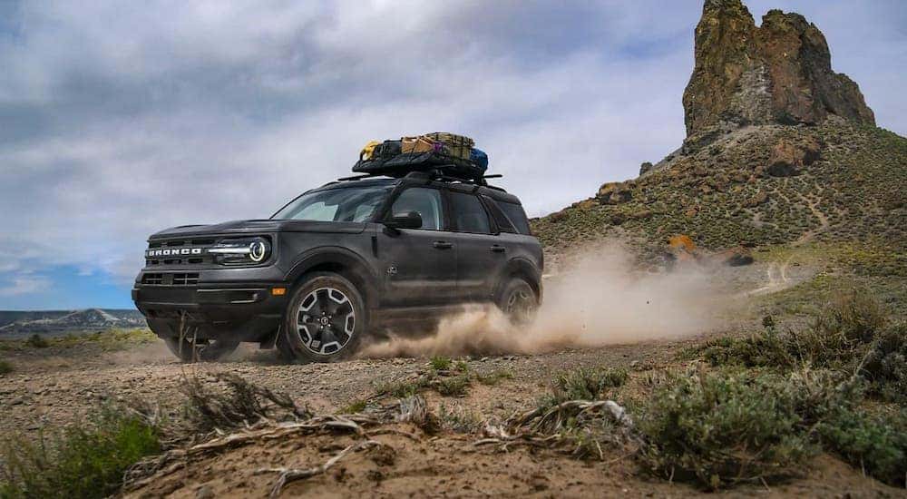 A black 2021 Ford Bronco Sport with luggage on the roof is driving on a dirt trail in front of rock formations.