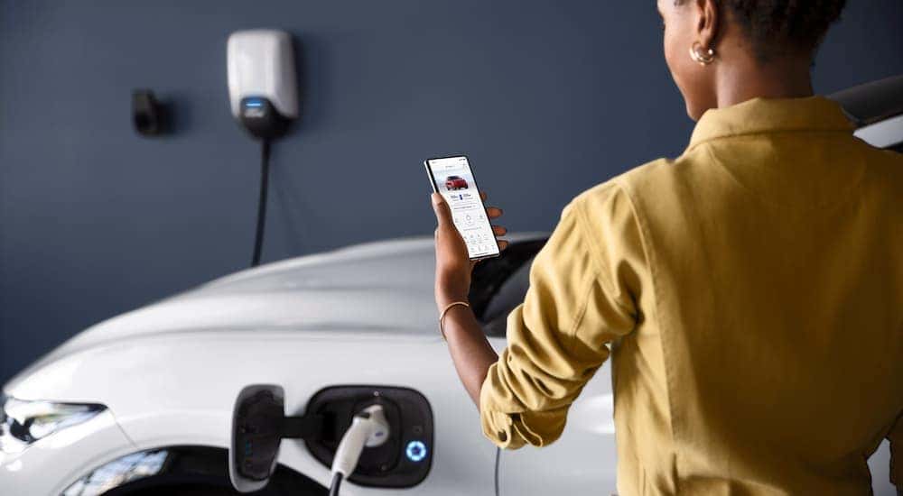 A woman is shown checking on a white 2022 Ford Mustang Mach-E through the Ford phone application as it charges.