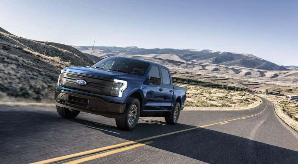 A blue 2022 Ford F-150 Lightning is shown from the front at an angle while it drives down a desert road.