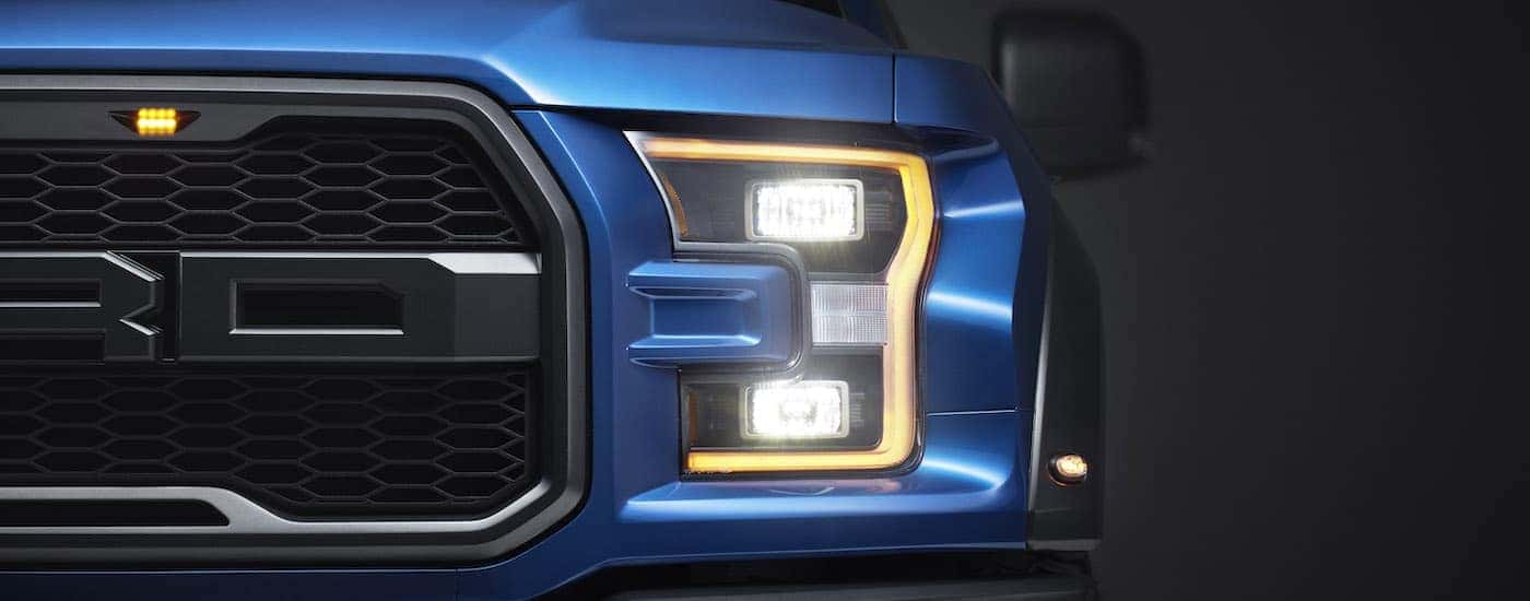 A close up shows the illuminated driver side headlight on a blue 2017 Ford F-150 Raptor.