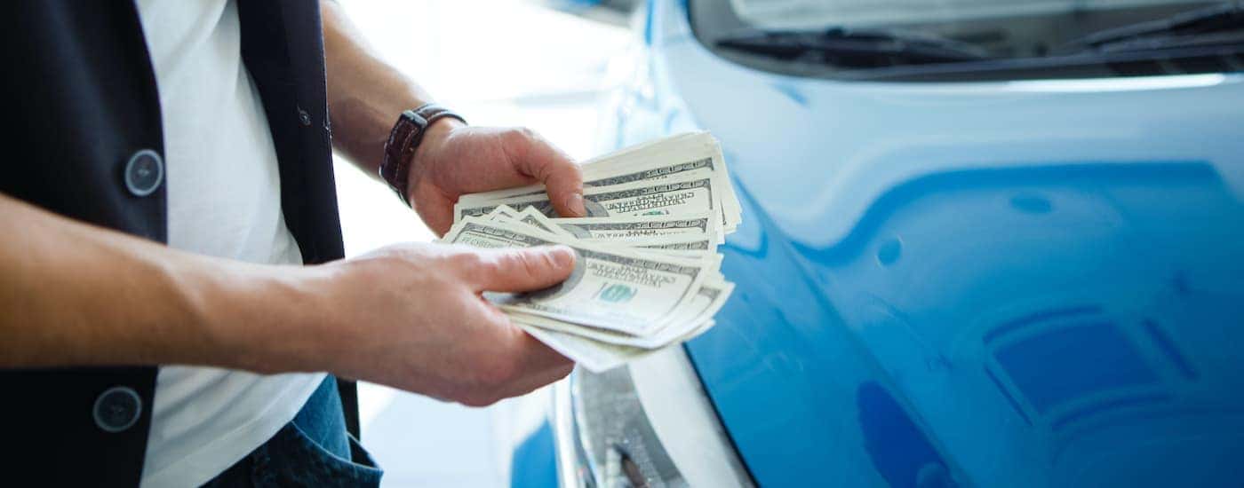 A close up shows a person holding cash in front of a blue car.