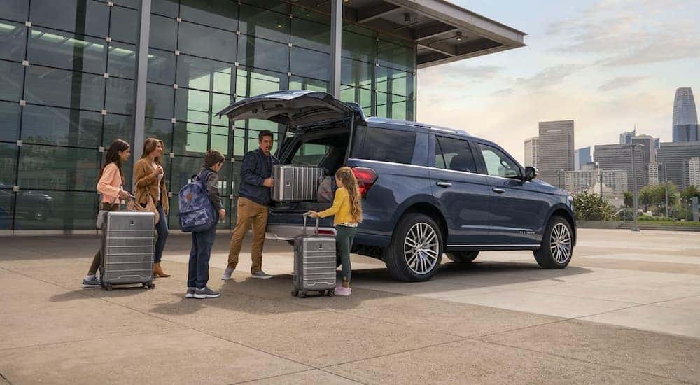 A family loads their matching luggage into the trunk of a blue 2023 Ford Expedition near a large glass building