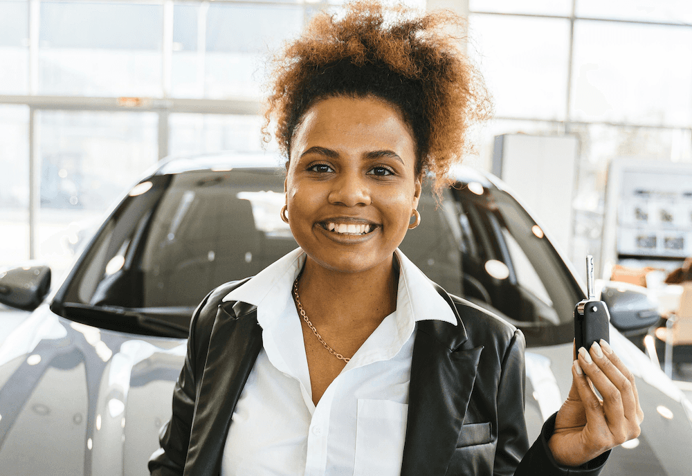 A smiling woman holds a car key and stands in front of the used car she bought in Richmond, VA