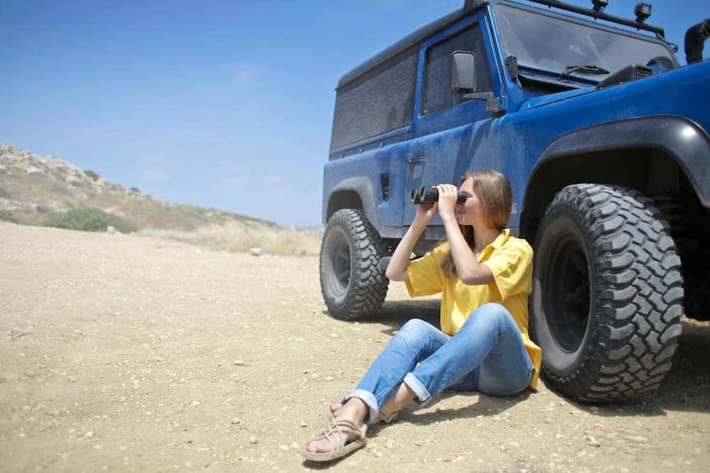 Woman on a summer road trip looking through binoculars sitting next to her Jeep SUV